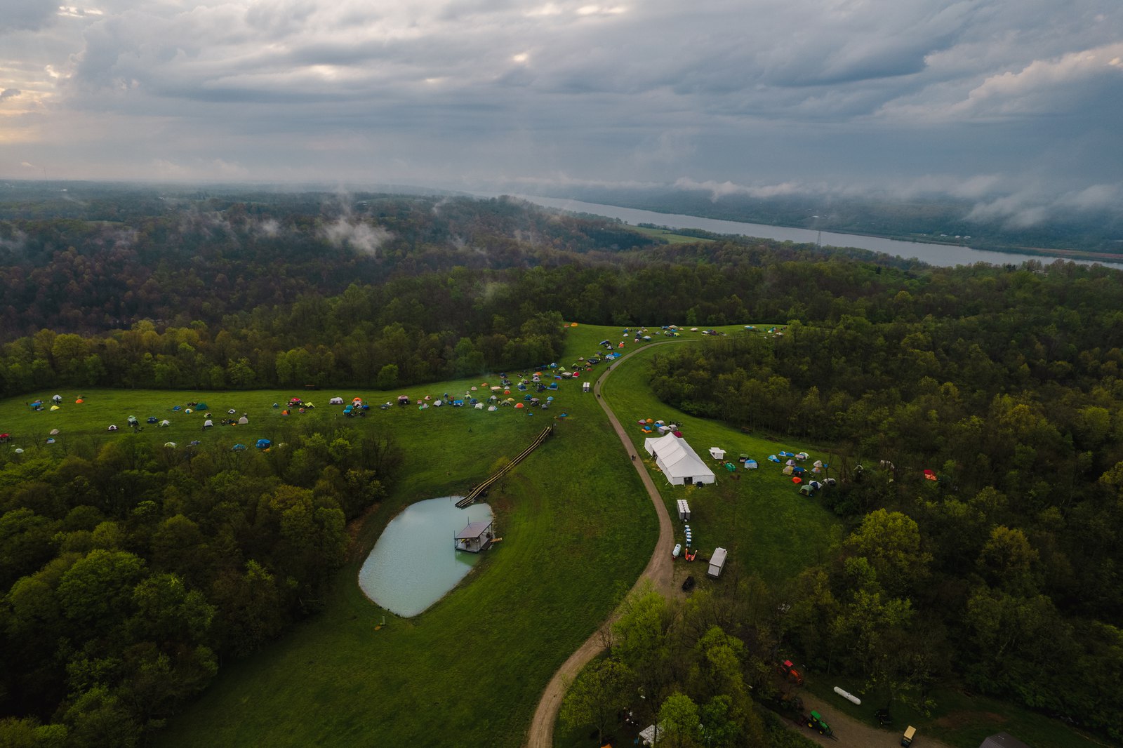 Tents, a pond and a white event tent in green hills under low clouds