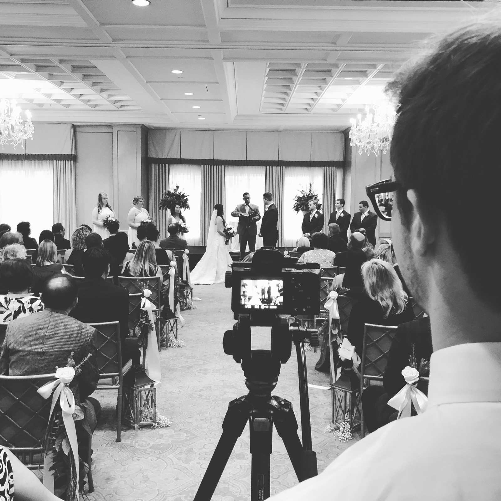 Black and white photo over Isaac's shoulder as he films a wedding ceremony on a tripod