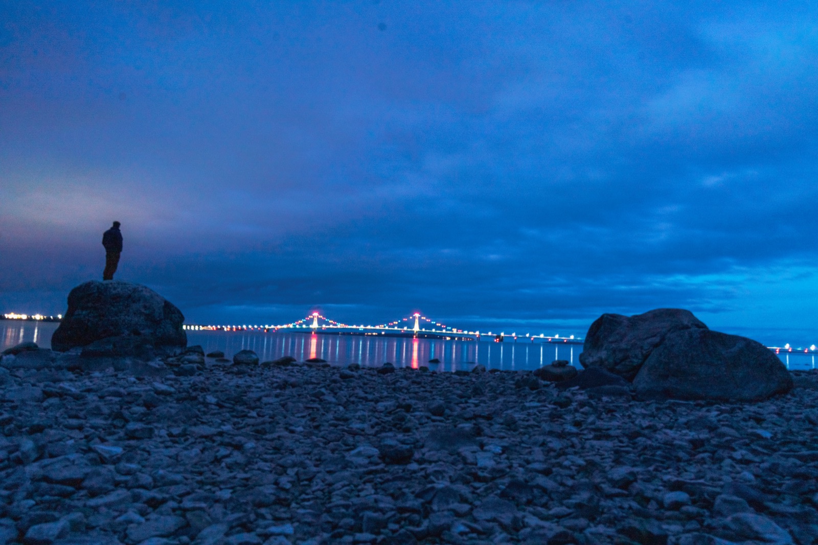 Man with a headlamp on rocks by a lit bridge at night
