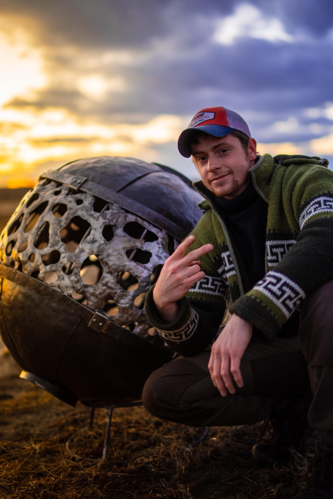 Man crouched beside a metal sphere sculpture at sunset